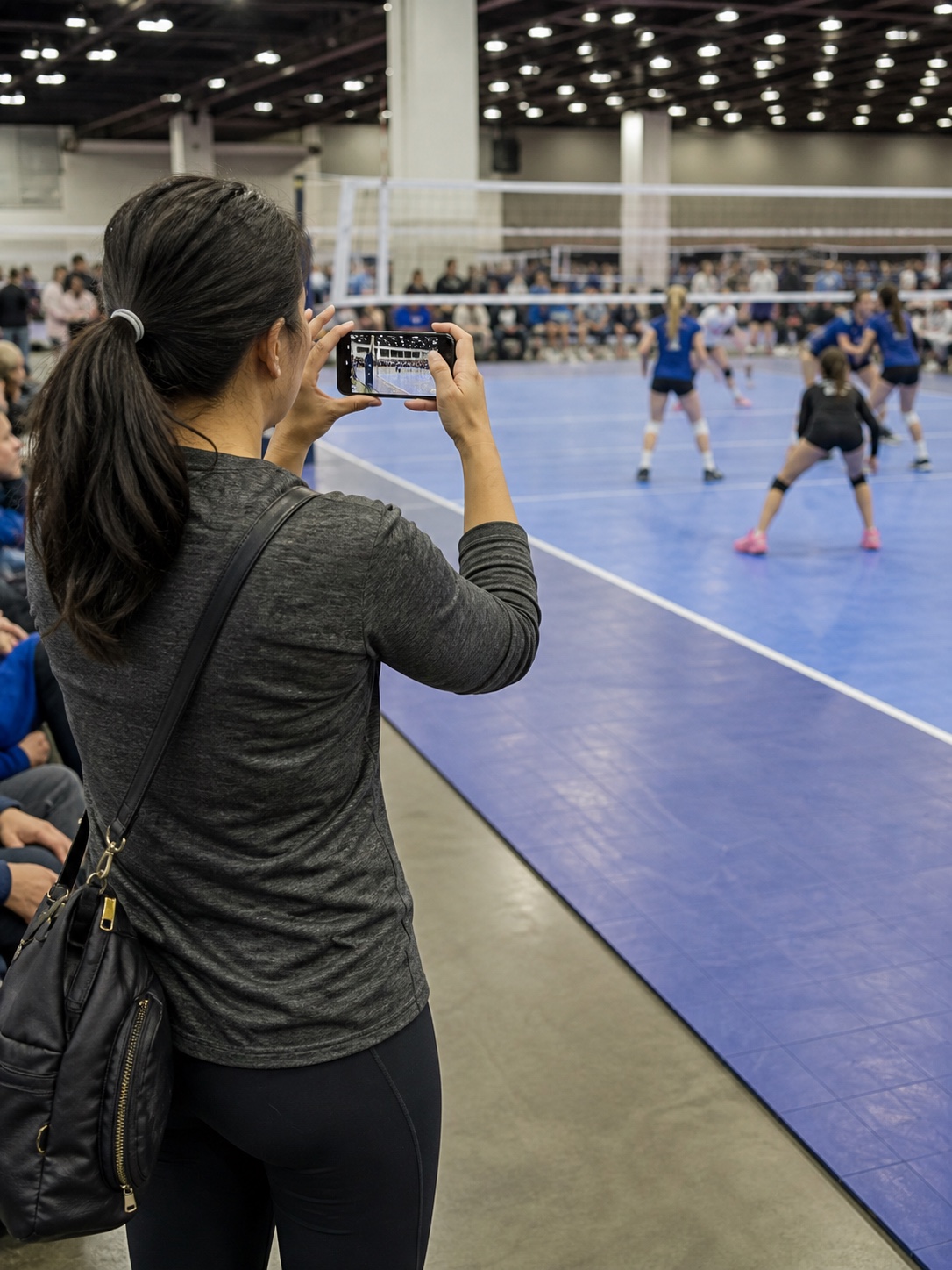 A parent recording a volleyball match with one phone in a crowded tournament gym